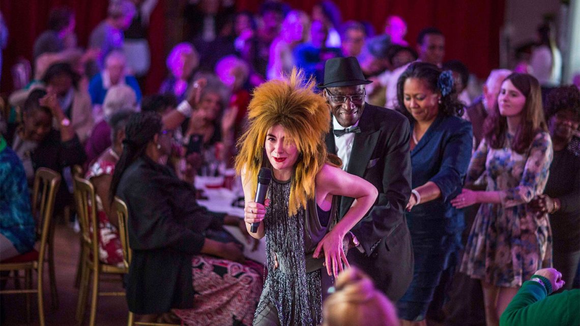 A performer in a big spiky wig at The Posh club leads a conga line of older adults.