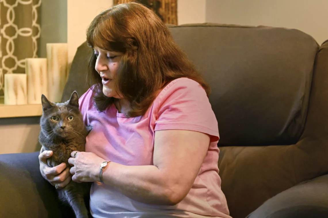 Cindy Tine, a white woman with brown hair wearing a pink t-shirt, is holding a large gray cat next to her.