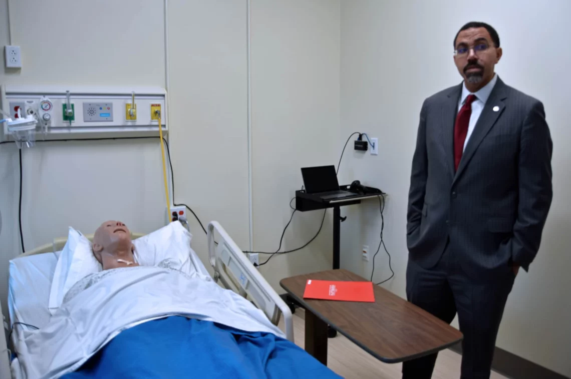 SUNY Chancellor John King is standing in a Nursing Skills Lab at SUNY Plattsburgh. John King is wearing a dark suit and has his hands in his pockets, he is standing in a hospital room where a practice mannequin is in the hospital bed in front of him.