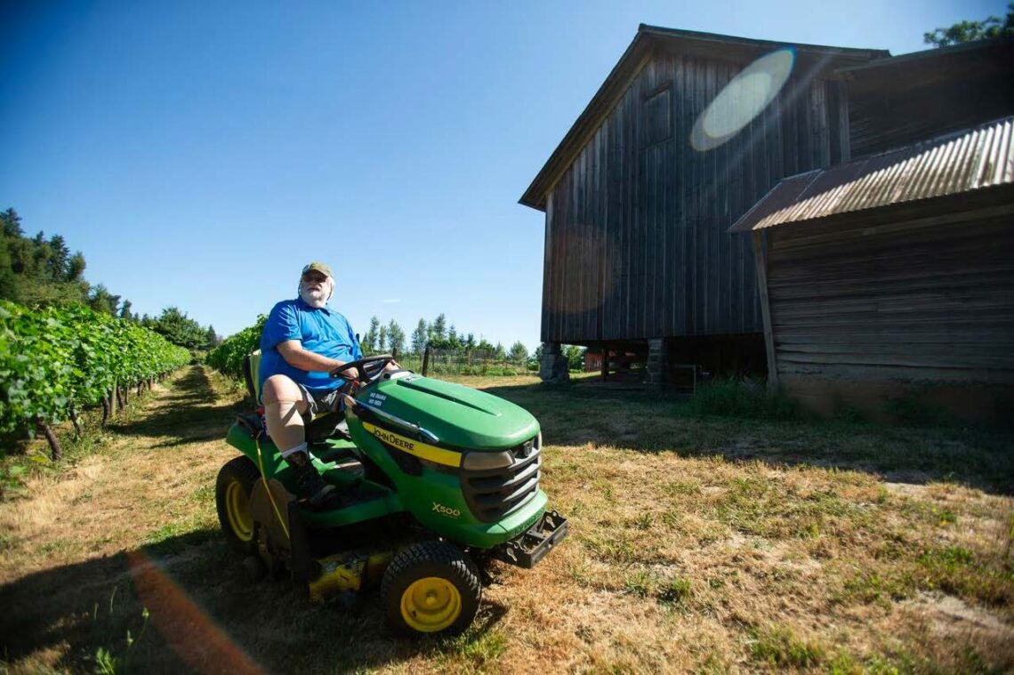 Farmer Rick Cook is a white man wearing a blue T-shirt and tan baseball cap. He is sitting on a green John Deere tractor in front of a barn on his Farmstead.
