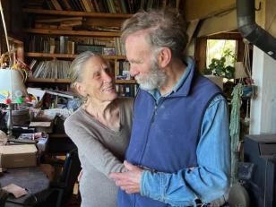 82-year-olds Jane Lapiner (left) and David Simpson, formerly known as the Diggers embrace in their living room, with a bookshelf in the background. He is wearing a blue shirt and darker blue vest, she is wearing a gray sweater.