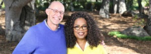 A man and a woman, both beaming smiles, stand close together outdoors in front of a tree on a sunny day. The man wears glasses and a blue sweater, while the woman, with curly hair and glasses, sports a yellow sweater. Their warmth reflects the bright spirit of Berkeley Journalism.
