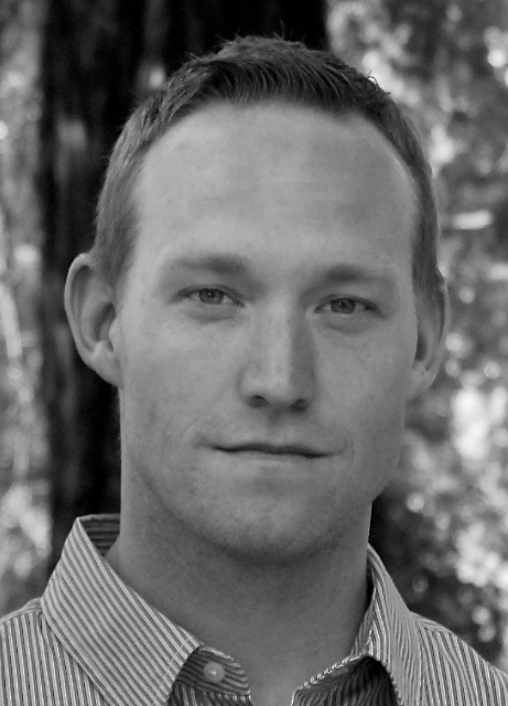 A black-and-white portrait of a person with short hair, wearing a striped collared shirt. The individual faces forward with a neutral expression, exuding an aura of inquisitiveness often seen in Berkeley Journalism alumni. The background is blurred, hinting at trees or foliage.