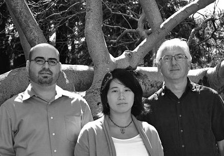 A black-and-white photo taken by a Berkeley Journalism student shows three people standing in front of a large, leafless tree. They are all gazing directly at the camera. Two men are on the left and right, and a woman is in the center. The background reveals branches and shadows from the tree.