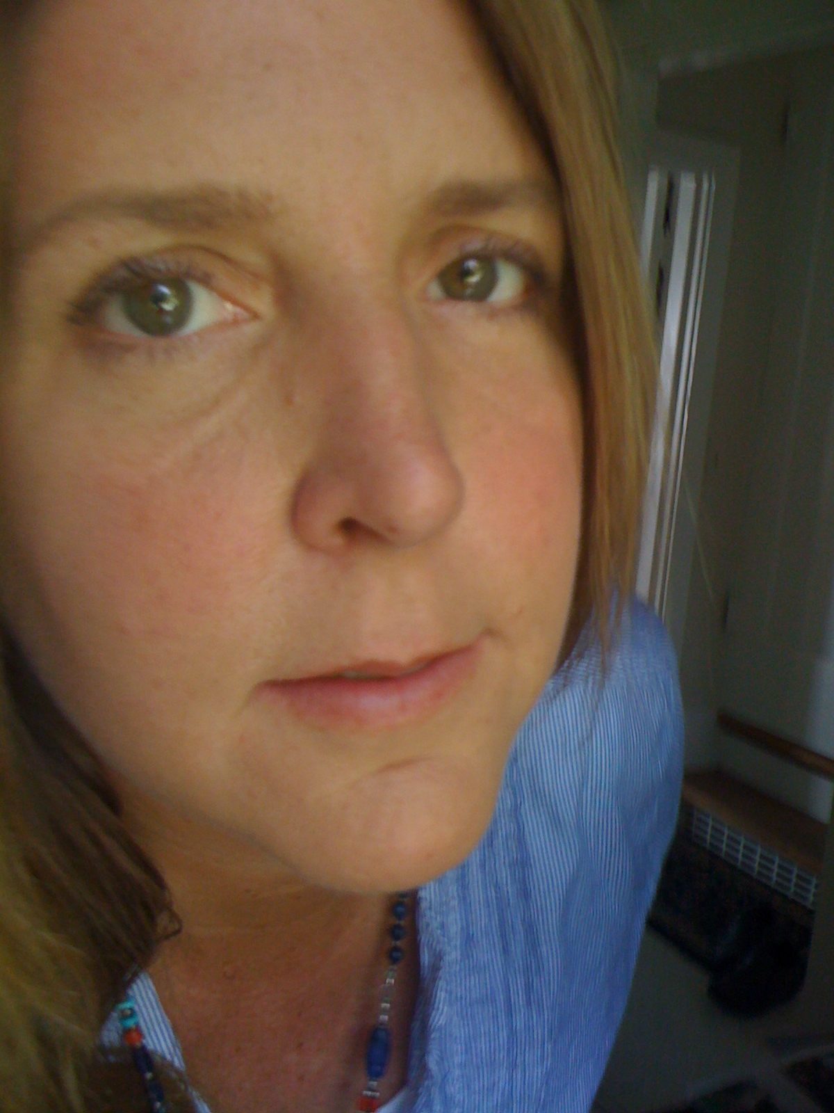 A close-up photo of a person with light brown hair and green eyes, slightly smiling. They are wearing a Berkeley Journalism blue shirt and a colorful beaded necklace. The background appears to be indoors with a glimpse of a door and some household objects.