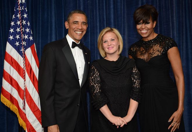 A man in a black tuxedo and a woman in a black gown stand on either side of Angela Hart '14, who is wearing a black dress and shawl. They are in front of a blue curtain with an American flag visible on the left. All three individuals, recipients of the White House Correspondents Association Scholarship, are smiling at the camera.