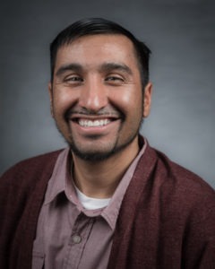 Juan Martinez, a man with short dark hair and a goatee, is smiling at the camera. He is wearing a maroon cardigan over a button-up shirt. The background is a plain, dark gray.