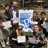A group of seven people, a mix of students and professional journalists, stands and kneels around a large sign with "SPJ" on it. They are outdoors on grass, holding awards certificates and smiling at the camera. Some are wearing casual clothes, while others are dressed in more formal attire.
