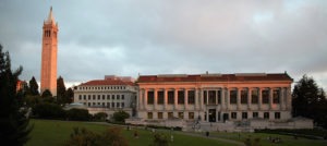 The image shows the UC Berkeley campus, with the prominent Sather Tower (Campanile) on the left and the grand Doe Library on the right. The buildings are illuminated by the sunset, with a green lawn and several people in the foreground, possibly including members of the Graduate School of Journalism Advisory Board.
