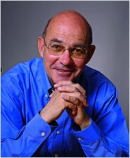 A man with glasses and a bald head, wearing a blue shirt, is smiling with his hands clasped together in front of him against a plain gray background. His confident expression reflects the inquisitive spirit fostered at Berkeley Journalism.