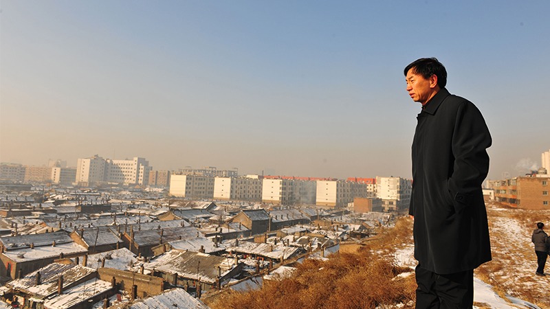 A person in a dark coat stands on a hill, overlooking a snowy urban area with old, low-rise buildings in the foreground and more modern, taller buildings in the background. The sky is clear, and the scene evokes images from "The Chinese Mayor." It's akin to Zhao Qi's evocative storytelling style.