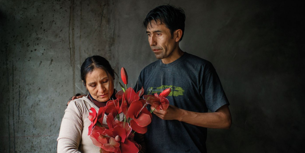 A man and woman stand close together against a dark backdrop. The woman, with downcast eyes, clutches a bouquet of red flowers. The man, reminiscent of a Daniel Alarcón character in his dark T-shirt, stands beside her with a comforting hand on her shoulder, looking thoughtfully ahead.