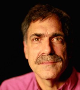 A man with brown hair, mustache, and beard stubble gazes at the camera with a serious expression. He is wearing a magenta shirt. The dark background adds to the intensity of David Tuller's contemplative look.