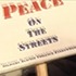 A small sign with the text "PEACE ON THE STREETS" written in large uppercase letters. The text is primarily in bold and black, with the word "PEACE" highlighted in red. The sign is attached to a wooden handle, symbolizing the collaborative efforts of TV students inspired by KQED.