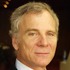 A man with short, light gray hair is looking directly at the camera. He has a light complexion and is wearing a dark-colored suit. The background is blurred and dark, suggesting an indoor setting. Ed Wasserman exudes confidence in his poised stance, possibly hinting at his recent Dean appointment.