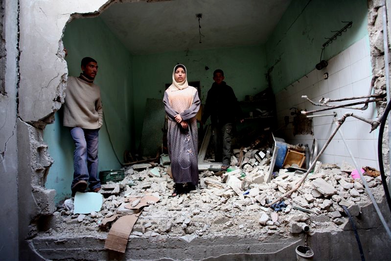 Three people stand in the middle of a demolished room with debris and rubble covering the floor. The walls are damaged, and broken tiles are visible. The central figure is a woman wearing a hijab, flanked by two men. A Berkeley Journalism team captures this scene amidst the chaos.