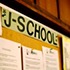 A close-up shot of a notice board with various documents pinned to it. A large sign reading "J-SCHOOL" is prominently displayed at the top of the board. The background is wooden, giving a warm tone to the scene. Among the papers, a highlighted notice announces that a prestigious grant has been awarded.