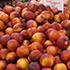 At a market, a large pile of ripe peaches with golden yellow and reddish hues is displayed. A sign in the background reads "Peaches $2.99 per pound." The slightly fuzzy texture of the peaches adds to their appeal as they are closely arranged together, reminiscent of a scene from Gabriela Quiros' PBS features.