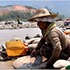 A person wearing a wide-brimmed hat and protective clothing works among a pile of large rocks in a rocky, arid riverbed. The background features a distant, hilly landscape under a blue sky—an ideal location for documenting environmental changes for an Internews project.