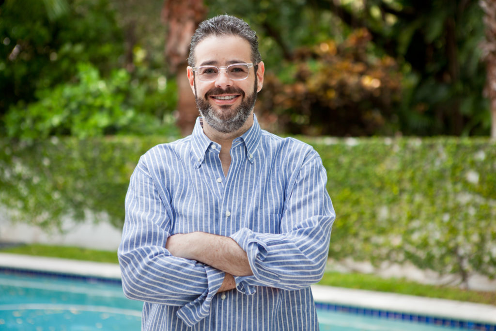 Isaac Lee, a man with a beard and glasses, wearing a blue-and-white striped shirt, stands outdoors with his arms crossed and smiles at the camera. The President of News at Univision is in front of a swimming pool with greenery in the background.