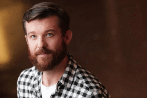 A man with a brown beard and mustache, wearing a black and white checkered shirt over a white t-shirt, looks directly at the camera. The background is blurred with a warm, brownish tone, reminiscent of an LA Times feature on virtual reality experiences.