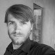 Black and white photo of a man with short hair and a beard, wearing a dark shirt. He is looking directly at the camera with a neutral expression. The background appears to be an indoor setting with a window and part of a wall visible. He recently participated in the New York Hackathon.