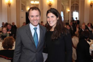 A man in a gray suit with a blue tie and a woman in a black dress, identified as Lauren Rosenfeld, smile for the camera at a formal event. The background shows a large room filled with seated people and ornate chandeliers, suggesting they are attending the Prestigious OPC Foundation Award celebration.