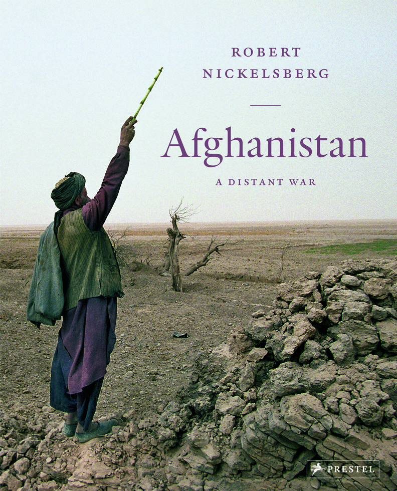 A person in traditional Afghan clothing stands in a barren, rugged landscape, raising a yellow stick toward the sky. The image is the cover of "Afghanistan: A Distant War" by Robert Nickelsberg, published by Prestel.
