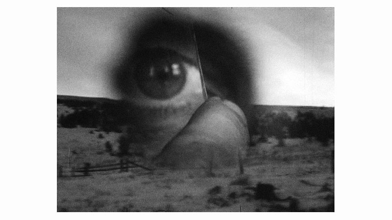 A surreal black and white image reminiscent of Robert Frank's films with a large translucent eye overlaying a barren landscape. The eye, magnified and centrally placed, seems to watch over the blurred scenery of what appears to be a desert or open plain. The image evokes a sense of mystery and observation.