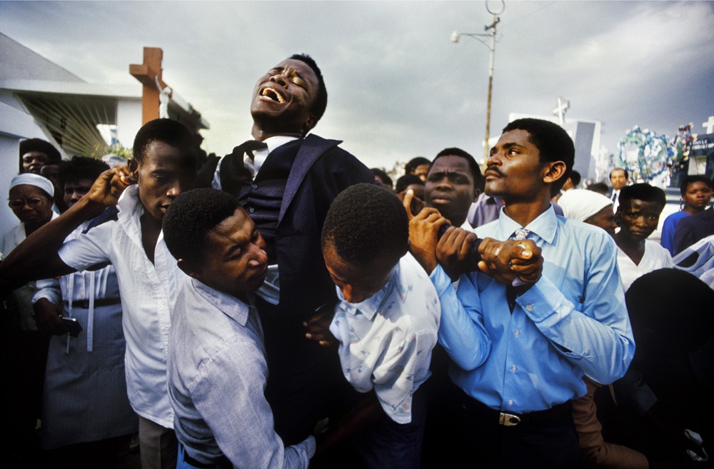 A grieving man in a suit is held and supported by several men and women, including nuns, in a crowded outdoor setting. The emotional scene captures a sense of loss and community support. Photographs from Haiti depict crosses and other people visible in the background under a cloudy sky.