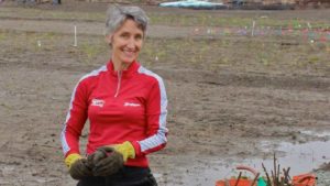 A smiling person with short gray hair and a red long-sleeve shirt is standing in a muddy field holding small plants. They are wearing yellow gloves. In the background, there are rows of small plants, colorful flags, and various work equipment. This former assistant dean is also a UC Berkeley Journalism alum from 1992.