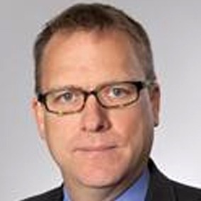 A man with short hair, wearing glasses, a blue dress shirt, and a dark suit jacket is looking directly at the camera. The background is a plain gray gradient. This image serves as a poignant moment in remembering Mark Hummels.