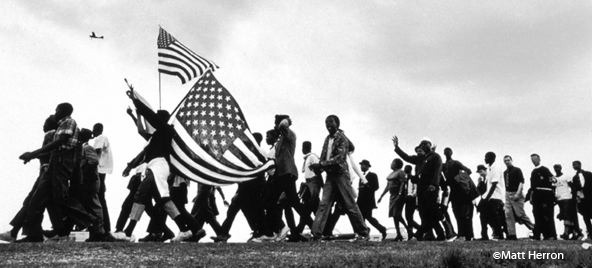 A black and white photo by Matt Herron depicts a large group of people, some holding American flags, marching with determined expressions. A plane is visible in the sky above. The image captures the essence of the Civil Rights movement.