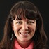 A woman with long brown hair, wearing a pink top and dangling earrings, smiles at the camera against a black background, reminiscent of a Dorothea Lange portrait.