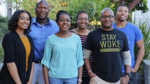 A portrait of six smiling people stands outdoors in a garden area with lush greenery in the background. They are dressed casually and appear friendly and relaxed. One person is wearing a "Stay Woke" shirt, possibly hinting at their connection to J-School or NABJ.