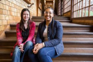 Two young women are sitting side by side on a wide wooden staircase inside a building. One is wearing a red sweater, while the other is in a black jacket. They are both smiling and looking at the camera. Large windows line the wall on the right side, reminiscent of Danielle Parenteau's student journalism for the New York Times.