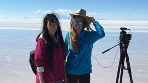 Two women, recent J-School graduates, stand on a vast salt flat under a clear blue sky. One wears a pink jacket and the other is in blue with a hat and sunglasses. A camera on a tripod stands beside them, capturing footage for their New Media Thesis Project against the expansive white salt flats and distant mountains.