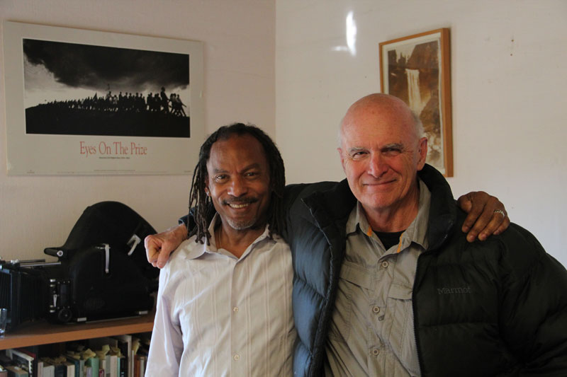 Two men smiling and standing side by side in a room. The man on the left has dreadlocks and wears a white shirt, while the man on the right has short hair and wears a gray shirt with a dark jacket. Behind them is a bookshelf, a framed picture, and a poster for 