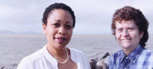 Two women stand outdoors near a body of water with a mountainous background. The woman on the left, Pamela Harris, has short curly black hair, wears a white blouse and a pearl necklace. The woman on the right has short curly brown hair, glasses, and a blue plaid shirt. They are both smiling like two film executives celebrating an Oscar nomination.