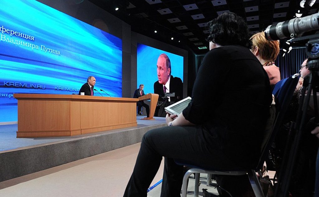 A man is speaking at a podium on a stage with a blue backdrop, while another man appears on a large screen behind him. Seated audience members, some taking notes, face the stage. The setting appears to be a formal press event discussing Putin's recent crackdown.