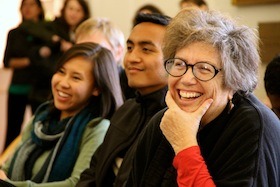 A group of people sit in a room, smiling and laughing. A woman with glasses and short hair in the foreground rests her chin on her hand, smiling widely. Nearby, a young man and woman, both grinning, are looking in the same direction, perhaps inspired by a story from their J-School days about Susan Rasky.