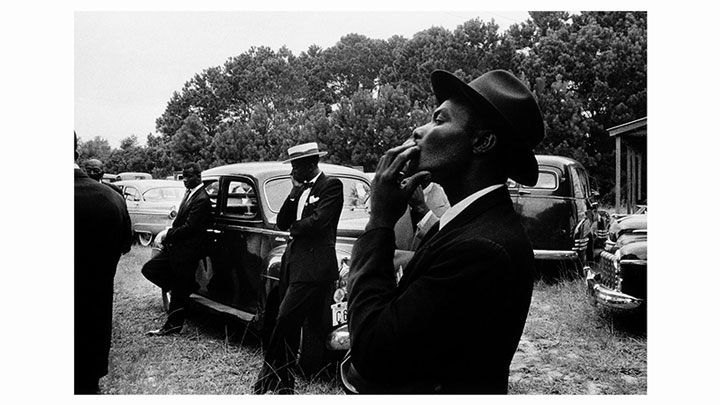 Black and white photo showing several men dressed in suits and hats in an outdoor setting. One man in the foreground is smoking a cigarette. Other men are leaning against vintage cars parked on grassy ground, with trees visible in the background—a snapshot of an era when analogue media reigned supreme.