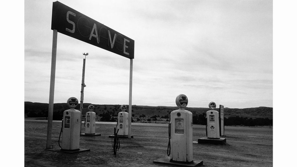 A black-and-white photo of an old, abandoned gas station with five vintage gas pumps scattered around. An overhead sign reads "SCA VE" against a cloudy sky. The scene, reminiscent of Logan's past, has a deserted, nostalgic feel with distant hills and sparse vegetation in the background.