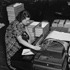 A woman operates a large machine in an office setting, surrounded by stacks of papers and folders. Dressed in a patterned dress, she is focused on the task at hand. The workspace, bustling with documents, reflects the dedication that earns National Winners the Mark of Excellence.