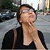A person with glasses and dark hair is standing on a city street, touching their chin with a thoughtful expression. They are wearing a black top and have a brown bag strap over their shoulder. In the background, buildings, cars, and trees frame the scene. It's almost as if Jessica Lum herself were pondering the secrets of SEO keywords.