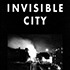 Black and white image with "INVISIBLE CITY" in bold white letters at the top. Below, a night view of a city street near the North Gate, with bright lights illuminating a foggy or smoky background, creating an eerie and mysterious atmosphere perfect for display.