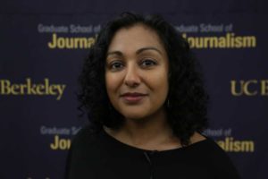 A person with shoulder-length curly hair stands in front of a backdrop that reads "Graduate School of Journalism" and "UC Berkeley." Tasneem Raja, wearing a black top, looks directly at the camera. The scene reflects the spirit of change at UC Berkeley's J-School.