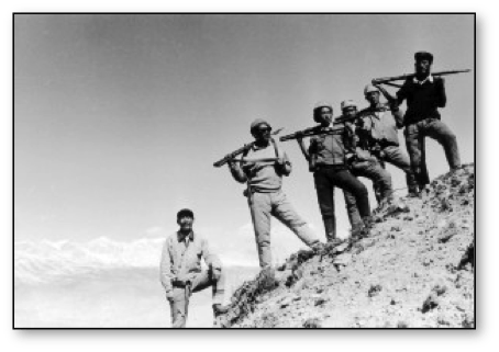 A black and white photo shows five people standing on a rocky slope. They are holding rifles on their shoulders, dressed in military-style uniforms and hats. The background features clear skies and what appears to be a mountain range in the distance, reminiscent of scenes from Ritu Sarin's Shadow Circus.