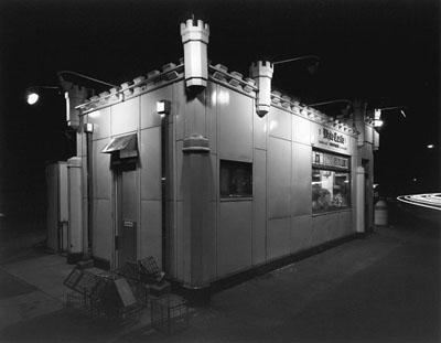 Black and white photo reminiscent of a George Tice piece, capturing a small, castle-themed building with turrets and crenellations. The large window reveals a bright interior while several crates rest near the entrance. Shrouded in minimal street lighting, this nocturnal scene evokes thoughts from the Fotovision Lecture.