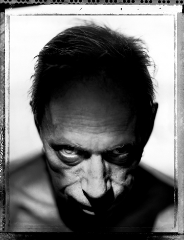 A black and white portrait photograph of Uncle Charlie with an intense and serious expression, looking directly at the camera from a low angle. His facial features are sharply defined due to the dramatic lighting, emphasizing deep shadows and highlights. Captured by the acclaimed photographer MARC ASNIN.